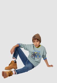 Maglione grigio chiaro con motivo di montagne, accenti blu, jeans blu, berretto marrone e stivali color tan con lacci gialli. Posizione seduta su uno sfondo neutro.