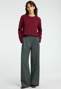 Burgunderfarbener Strickpullover mit Rundhalsausschnitt, kombiniert mit hochgeschnittenen, weiten grauen Nadelstreifenhosen. Glänzende braune Schuhe runden das Outfit ab.