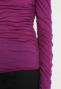 Lila Langarmoberteil mit geraffter Textur, seitlichen und Ärmeln mit Rüschendetails, gefertigt aus weichem, dehnbarem Material.