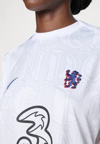Wit shirt met een verhoogd patroon van tekst en een prominent zwart grafiek, verfraaid met een geborduurd blauw en rood leeuwembleem op de borst.