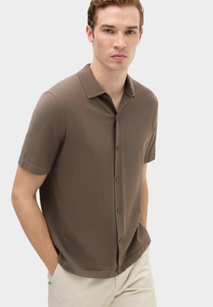 Junger Mann trägt ein braunes kurzärmeliges Hemd mit Kragen und beige Hose, steht mit den Händen in den Taschen.
