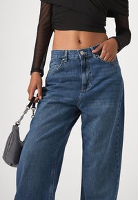 Szerokie niebieskie dżinsy z wysokim stanem, z klasycznymi kieszeniami i subtelną, wyblakłą fakturą. W zestawieniu z małą czarną torebką o teksturowanej powierzchni.