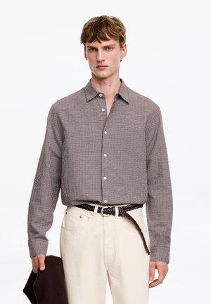 Jeune homme portant une chemise à carreaux marron rentrée dans un pantalon blanc avec une ceinture marron tressée, tenant une veste marron dans la main gauche, debout sur fond blanc.