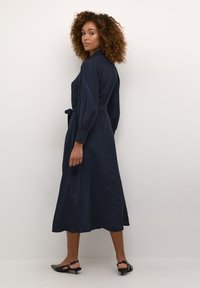 Marinblå knälång klänning med bälte i midjan, långa puffärmar och slät textur. Avslutad med en sid slits, bärs med svarta kitten heels.
