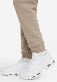 Bílé atletické tenisky s texturovaným syntetickým svrškem, vzdušným polstrováním v podrážce a jemnými šedými detaily, v kombinaci s béžovými tepláky.