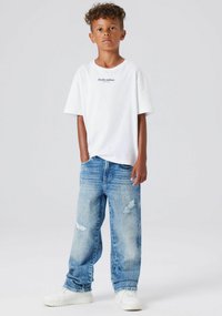 Maglietta bianca a maniche corte con scritta "Studio edition", abbinata a jeans di denim chiaro strappati e sneakers bianche.