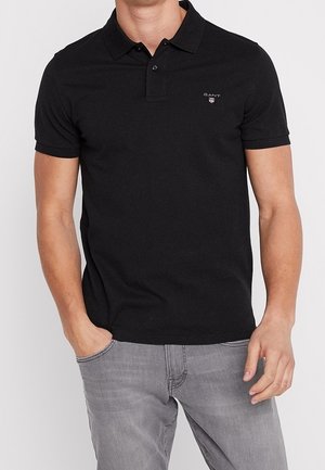 Homme portant un polo noir à manches courtes avec un petit logo GANT, associé à un jean gris clair, debout avec une main dans la poche.