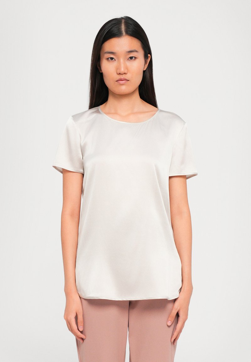Donna con lunghi capelli scuri che indossa una blusa di satin beige chiaro a maniche corte e pantaloni rosa tenui, in piedi contro uno sfondo neutro.