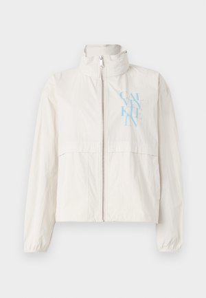 Lichte, crèmekleurige jas van een soepele stof met een hoge kraag, een ritssluiting aan de voorkant en een blauw logo op de linkerkant van de borst.