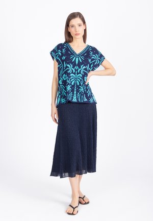 Kobieta ubrana w granatową bluzkę w kwiaty z dekoltem w serek, teksturowaną granatową spódnicę midi oraz czarne sandały, stojąca z jedną ręką na biodrze.