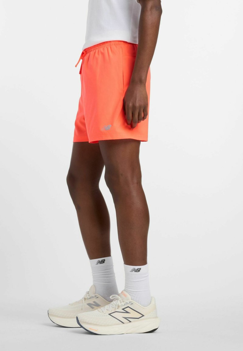 Shorts orange néon avec une taille élastique et un cordon de serrage. Présente un petit logo argenté sur l'ourlet. Portés avec des chaussures New Balance blanches.