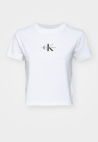 Bílý krátký rukávový top s cropped střihem, na hrudi s černým a šedým potiskem loga "cK Calvin Klein Jeans", kulatý výstřih, jednobarevné pozadí.