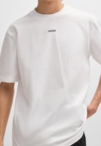 Biała bawełniana koszulka z okrągłym dekoltem i krótkimi rękawami; posiada małe, czarne logo "HUGO" na klatce piersiowej. Gładka faktura.