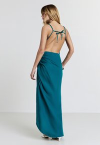 Frau mit blonden Haaren, die ein türkisfarbenes, rückenfreies Kleid mit dünnen Trägern trägt, das am Rücken gebunden ist. Sie steht und schaut nach links vor einem schlichten Hintergrund.