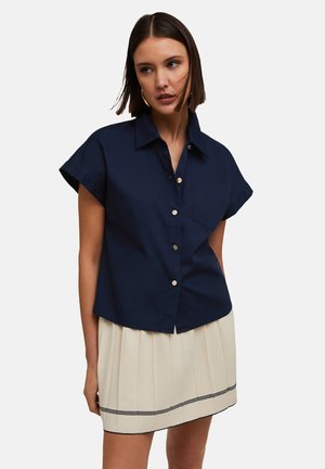 Camicia blu navy con maniche corte, tasca frontale e bottoni color oro, abbinata a una gonna plissettata crema con una striscia nera.