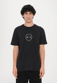 Sort t-shirt lavet af bomuld, med et rundt logo med "A|X" i hvidt i midten. Korte ærmer og rund halsudskæring.