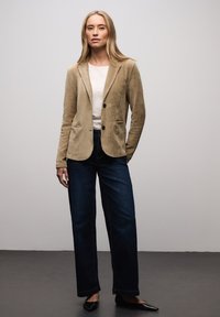 Blazer beige en velours côtelé avec col à revers, deux poches avant et boutons noirs, associé à un jean large en denim foncé et des chaussures noires.