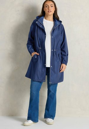 Mujer vestida con un impermeable azul marino con capucha, una camisa blanca, jeans azules y zapatillas blancas, de pie contra un fondo liso.