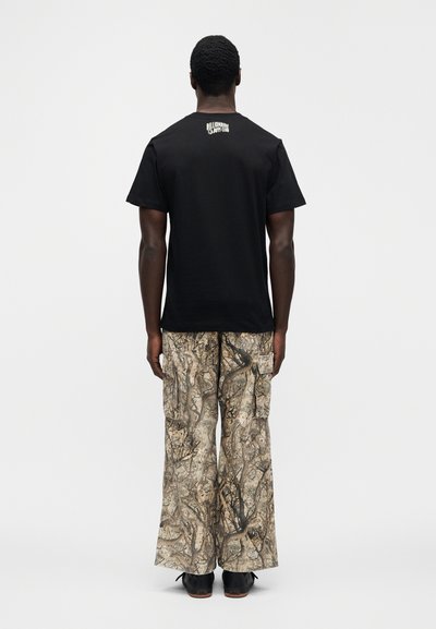 Μαύρο βαμβακερό μπλουζάκι με λογότυπο στην πλάτη, σε συνδυασμό με φαρδιά παντελόνια cargo με σχέδιο παραλλαγής σε μπεζ και καφέ αποχρώσεις.