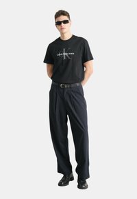 Camiseta de algodón negra con el logo "Calvin Klein Jeans", combinada con pantalones azul marino a rayas. Accesorios incluyen un cinturón negro y gafas de sol.