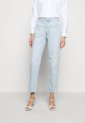 Vrouw draagt lichtblauwe high-waisted rechte jeans met schuine voorzakken, witte blouse met knopen en witte sandalen met hak.