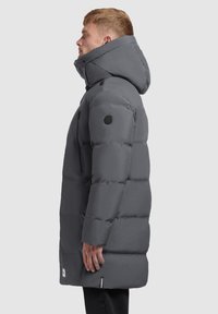 Muškarac stoji bočno i nosi dugu sivo puf jaknu s kapuljačom, zatvorenim džepom na prsima i crnim kružnim logom na rukavu.