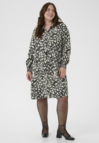 Vrouw die glimlacht, gekleed in een zwart-wit gestreepte lange mouw jurk, doorschijnende zwarte panty’s en zwarte hoge hakken Mary Jane schoenen, staand op een witte achtergrond.