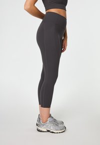 Leggings negros de tiro alto y ajustados, con paneles laterales texturizados, complementados por zapatillas deportivas grises con acentos reflectantes.