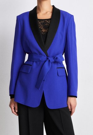 Frau trägt einen taillierten königsblauen Blazer mit schwarzen Revers, gebundenem Taillengürtel, Pattentaschen, über einem schwarzen Spitzenoberteil und schwarzen Hosen.