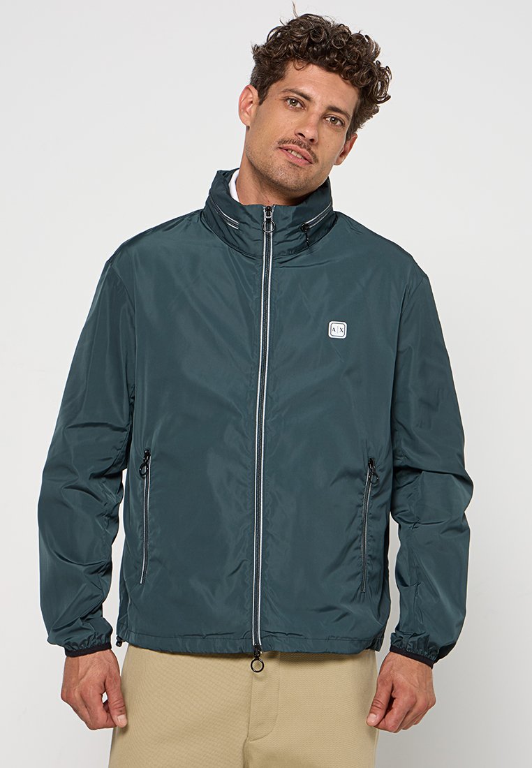Uomo con capelli ricci che indossa una giacca verde scuro con zip e tasche laterali e pantaloni beige, in piedi davanti a uno sfondo chiaro e uniforme.