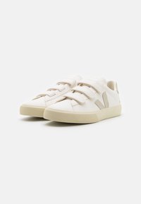 Baskets en cuir blanc avec des bandes Velcro, une accent beige sur le côté et une semelle en caoutchouc texturé. Design minimaliste avec une forme de chaussure à bout rond.
