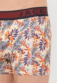 Herren-Badehosen mit einem mehrfarbigen Blumenmuster auf einem cremefarbenen Hintergrund, ausgestattet mit einem schwarzen Bund, auf dem "JACK&" in roter Schrift steht.