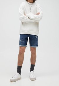 Sweat-shirt blanc, short en denim avec ourlet roulé, chaussettes grises et baskets blanches. Les caractéristiques notables incluent un effet usé sur le short et des colliers superposés.