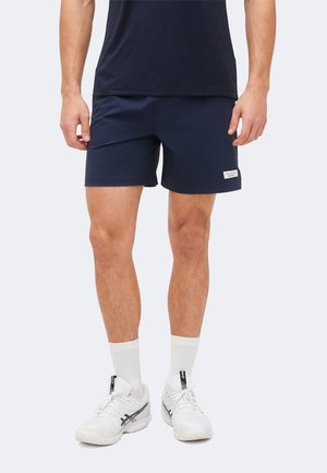 Muž nosící tmavě modré sportovní šortky, bílé kotníkové ponožky a bílé běžecké boty na jednobarevném světlém pozadí.