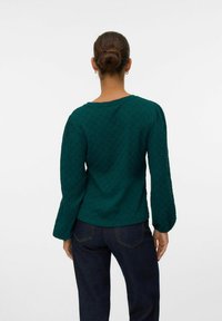 Mörkgrön texturerad topp med långa puffärmar, en båtformad halsringning och en något åtsittande silhuett, i kombination med mörka jeans.