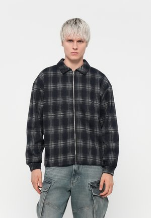 Black and gray plaid zip-up jacket with a collar, oversized fit, and ribbed cuffs, paired with light gray cargo pants featuring side pockets.