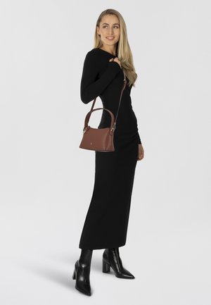 Donna con un lungo abito nero che indossa stivali neri con tacco, porta una borsa in pelle marrone con tracolla e manici.