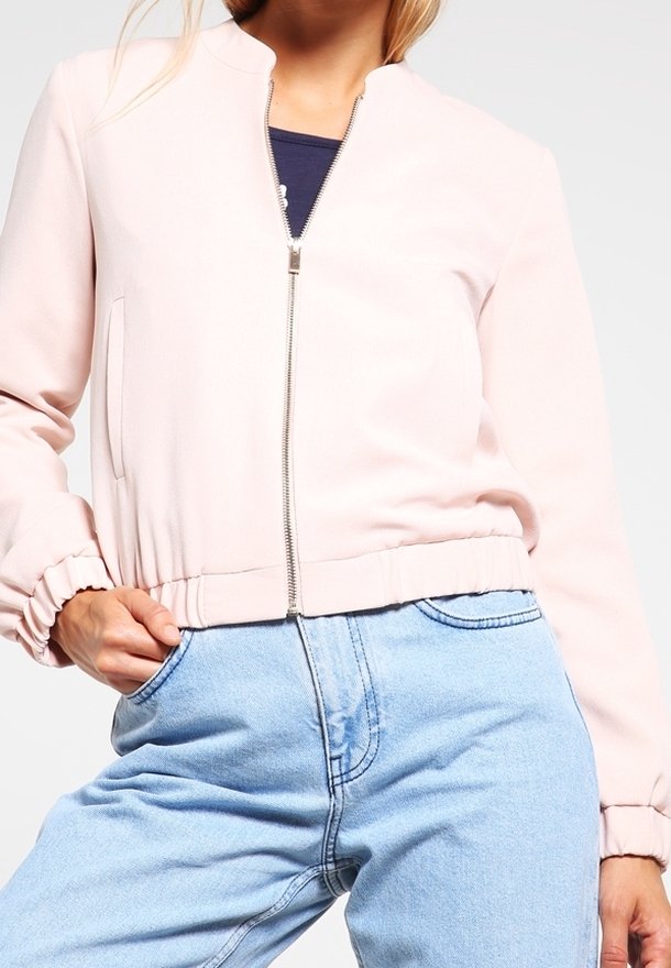Femme portant une veste zippée rose clair avec des poignets et un ourlet élastiques, associée à un jean léger bleu clair taille haute, se tenant avec une main dans la poche.