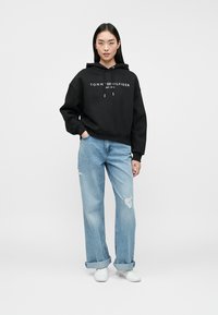 Frau steht in einem schwarzen Tommy Hilfiger Hoodie, light ripped weit geschnittenen Jeans und weißen Sneakers vor einem einfarbigen Hintergrund.