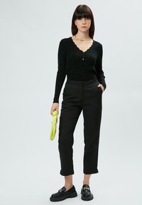 Schwarzes geripptes Langarmoberteil mit gewelltem Ausschnitt, kombiniert mit schwarzen maßgeschneiderten Hosen. Hält einen lim green Regenschirm und trägt schwarze Loafers.