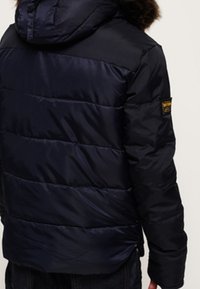 Giacca puffer blu navy con cappuccio, caratterizzata da cuciture orizzontali, colletto foderato in pelliccia e una toppa sulla manica sinistra. Texture liscia e lucida.