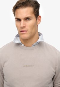 Lys grå sweatshirt med ribbet tekstur, prydet med et broderet "SUPERDRY" logo. En krave skjorte med blå og hvide striber ses tydeligt under.