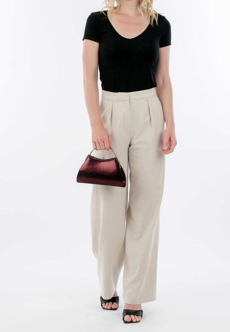 Femme portant un haut noir à manches courtes et un pantalon beige à jambes larges, tenant un petit sac à main rouge foncé brillant avec une poignée métallique.