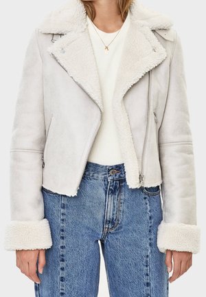 Femme portant une veste en peau retournée gris clair avec des manches retroussées, un haut blanc côtelé, un jean bleu taille haute et un collier pendentif en or.