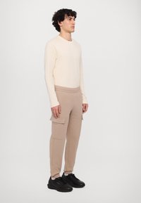 Beige langermet skjorte kombinert med lysebrune cargobukser med sidelommer. Svarte sko med teksturert overflate fullfører antrekket.