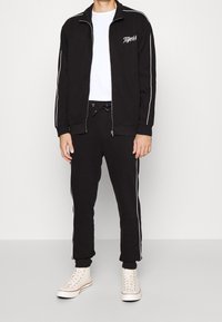 Veste de survêtement noire zippée avec des rayures blanches, associée à un pantalon de jogging noir assorti doté d'une taille à cordon, et des baskets montantes blanches.