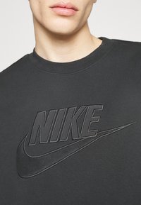 Detaljbild av en person som bär en svart sweatshirt med rund halsringning med en texturerad svart Nike-logotyp och swoosh på bröstet.