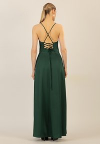 Langes grünes Satin-Kleid mit einem gekreuzten Rückendesign, Spaghettiträgern und einer gebundenen Akzentuierung in der Taille. Glatte Textur überall.