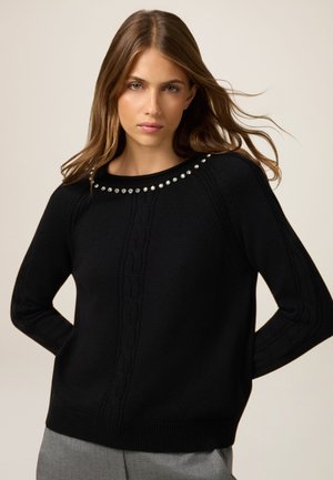Schwarzer Strickpullover mit einem gerippten Rundhalsausschnitt, geschmückt mit dekorativen silbernen Nieten. Enthält Zopfmusterdetails an den Seiten.