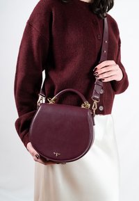 Okrągła, głęboka bordowa skórzana torebka z górnym uchwytem i regulowanym paskiem, wyposażona w złote okucia i małe logo.